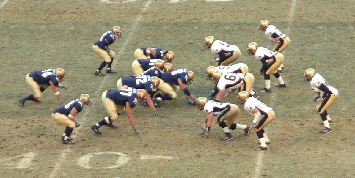 Football players set at the line of scrimmage before a snap, used as a visual anchor for draft-room decision-making under constraints.